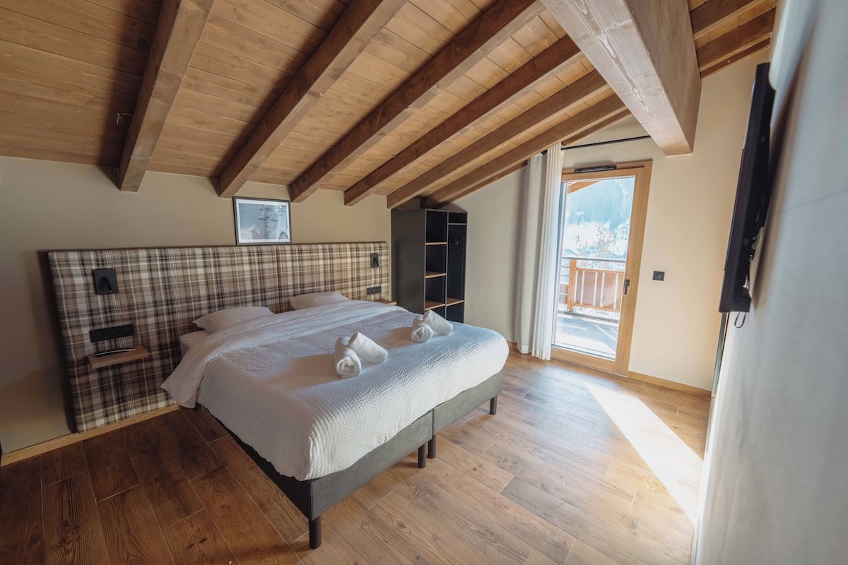 A bedroom with a bed, wooden beams, a plaid headboard, a small shelf, and a view of the outdoors.