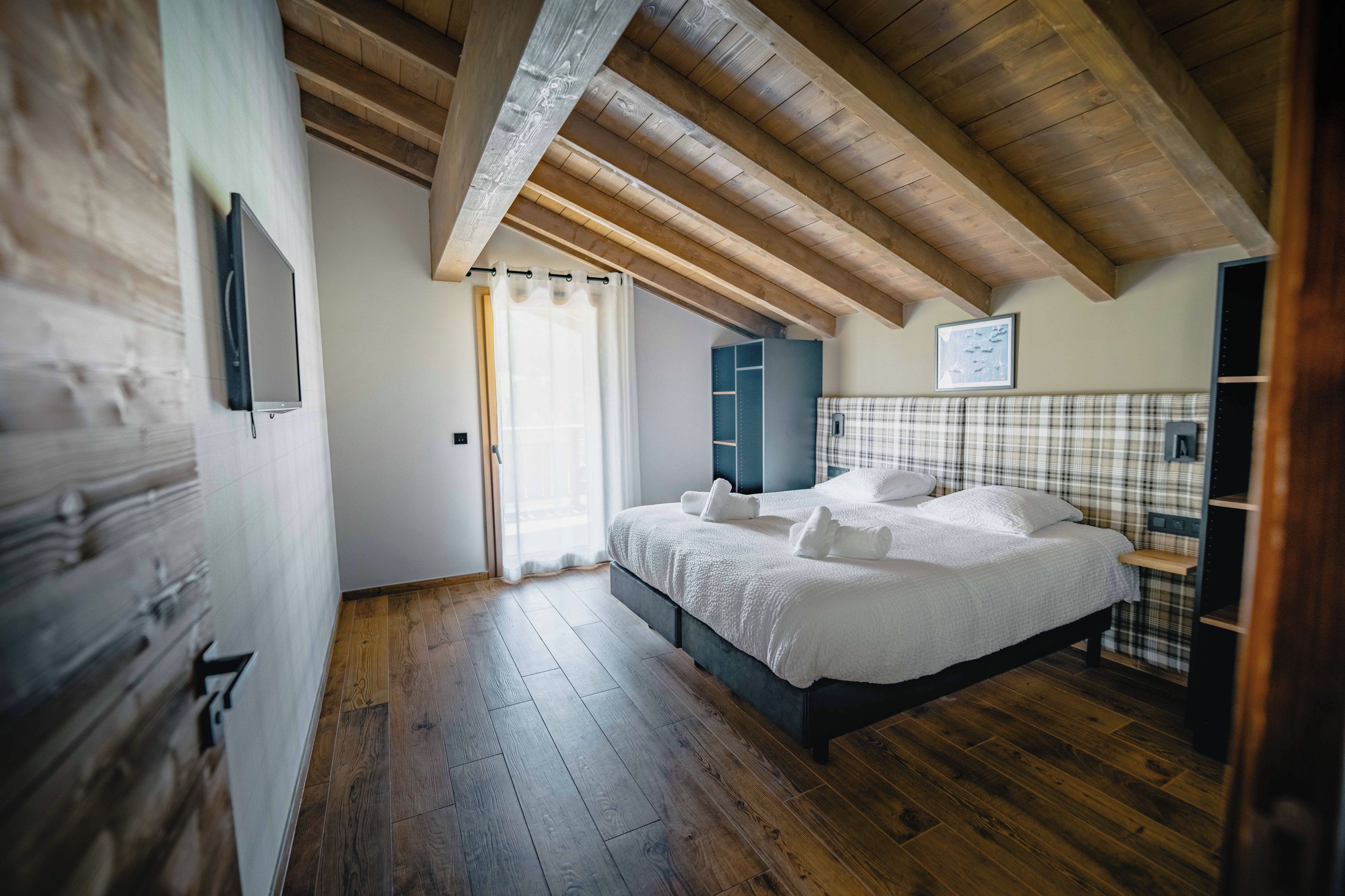 Une chambre avec un plafond en bois, un lit avec une literie blanche, une tête de lit à carreaux et une fenêtre ornée de rideaux légers.