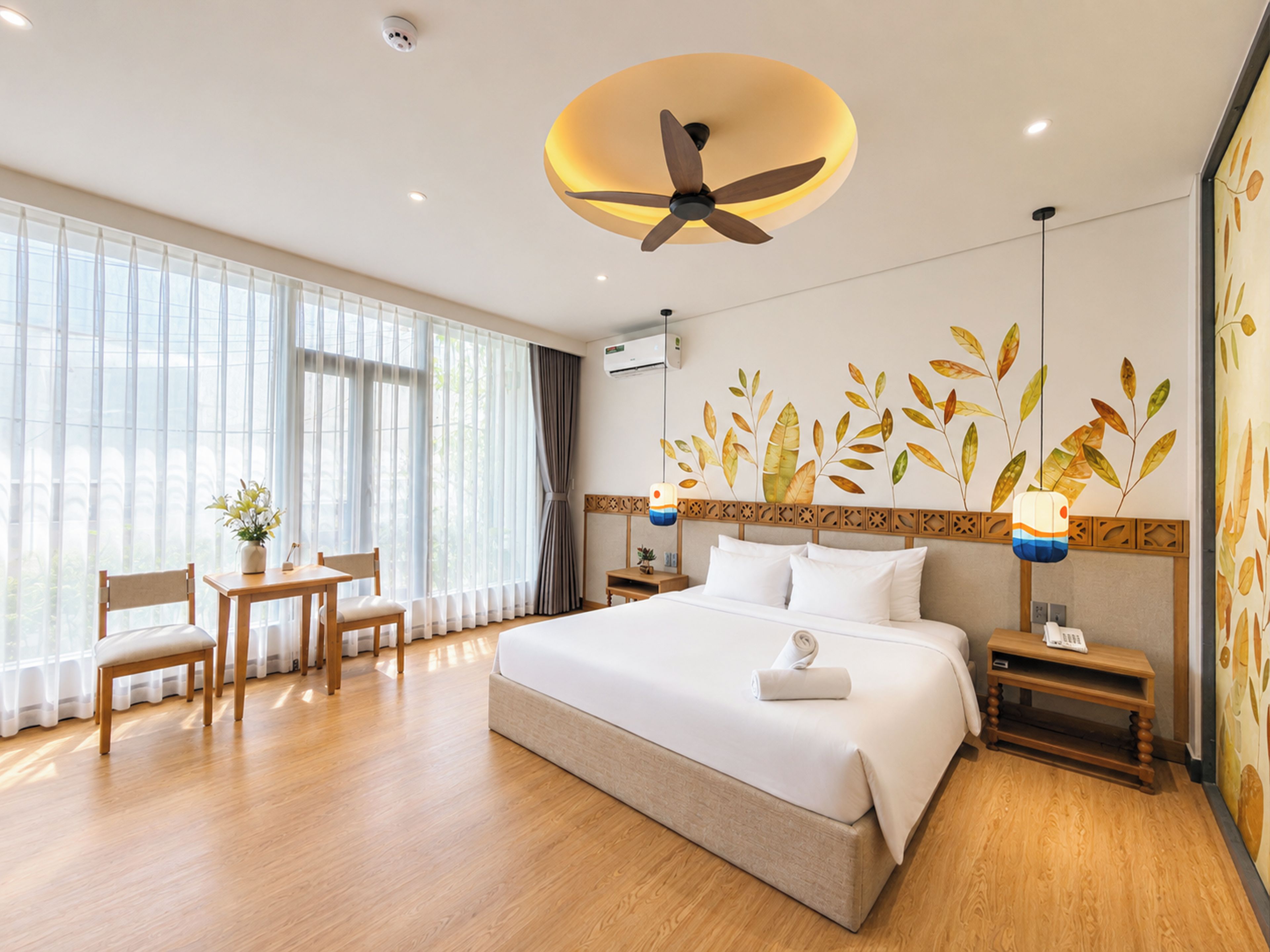 A hotel room with a large bed, a desk with a chair, a ceiling fan, and a wall with leaf patterns.