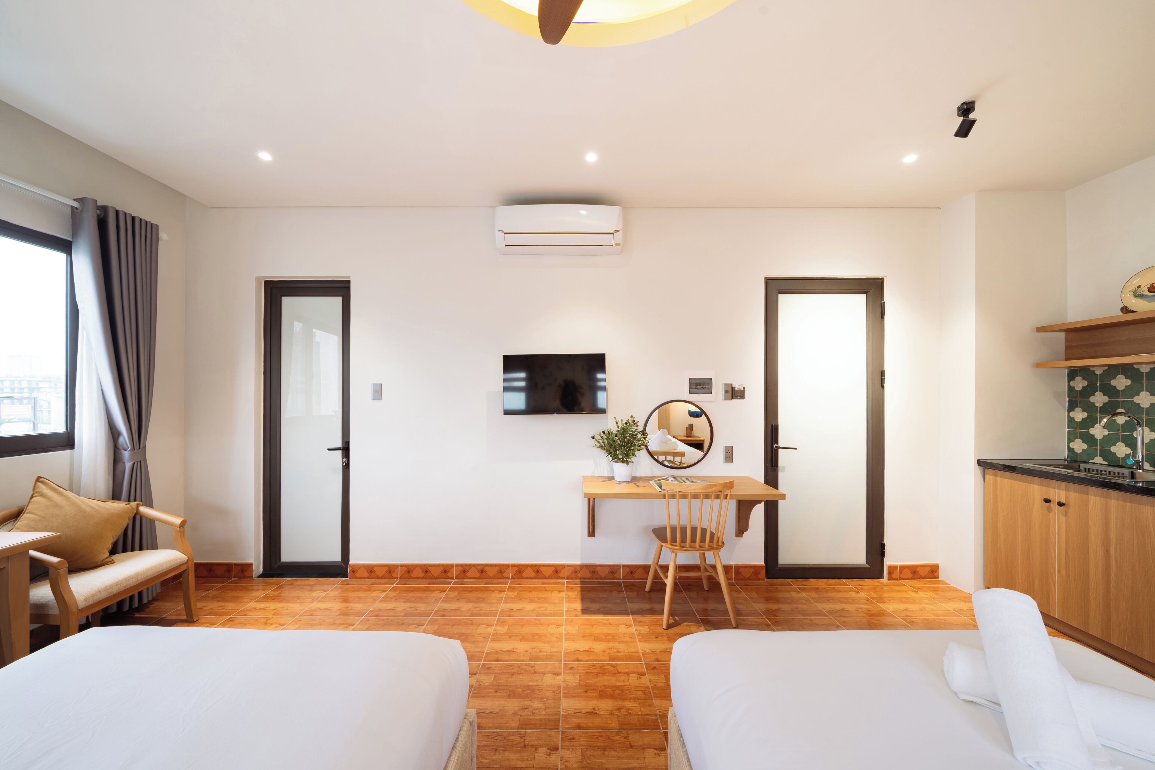 A hotel room with two beds, a desk with a chair, a TV, and a small kitchen area.