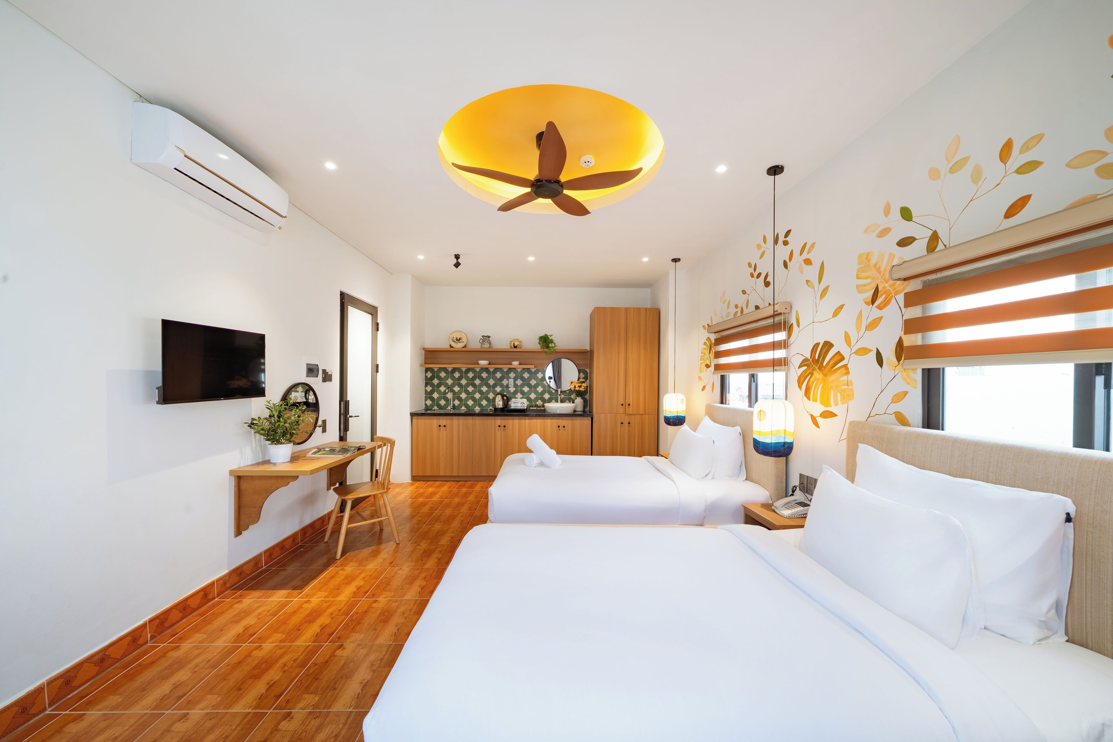 A hotel room with two beds, a desk, a TV, a ceiling fan, and a kitchen area in the background.