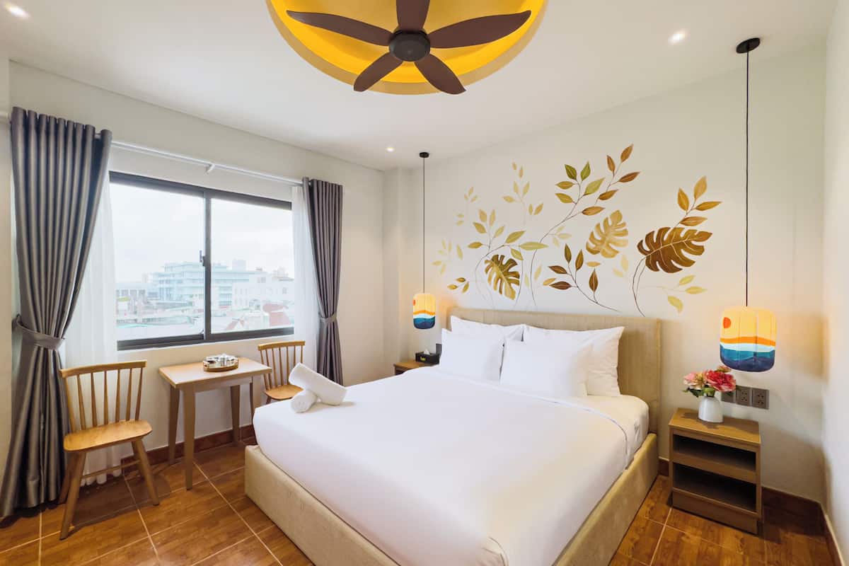 Habitación de hotel con cama, escritorio, silla, ventana con cortinas, ventilador de techo, decoración en la pared y mesita de noche con un jarrón de flores.