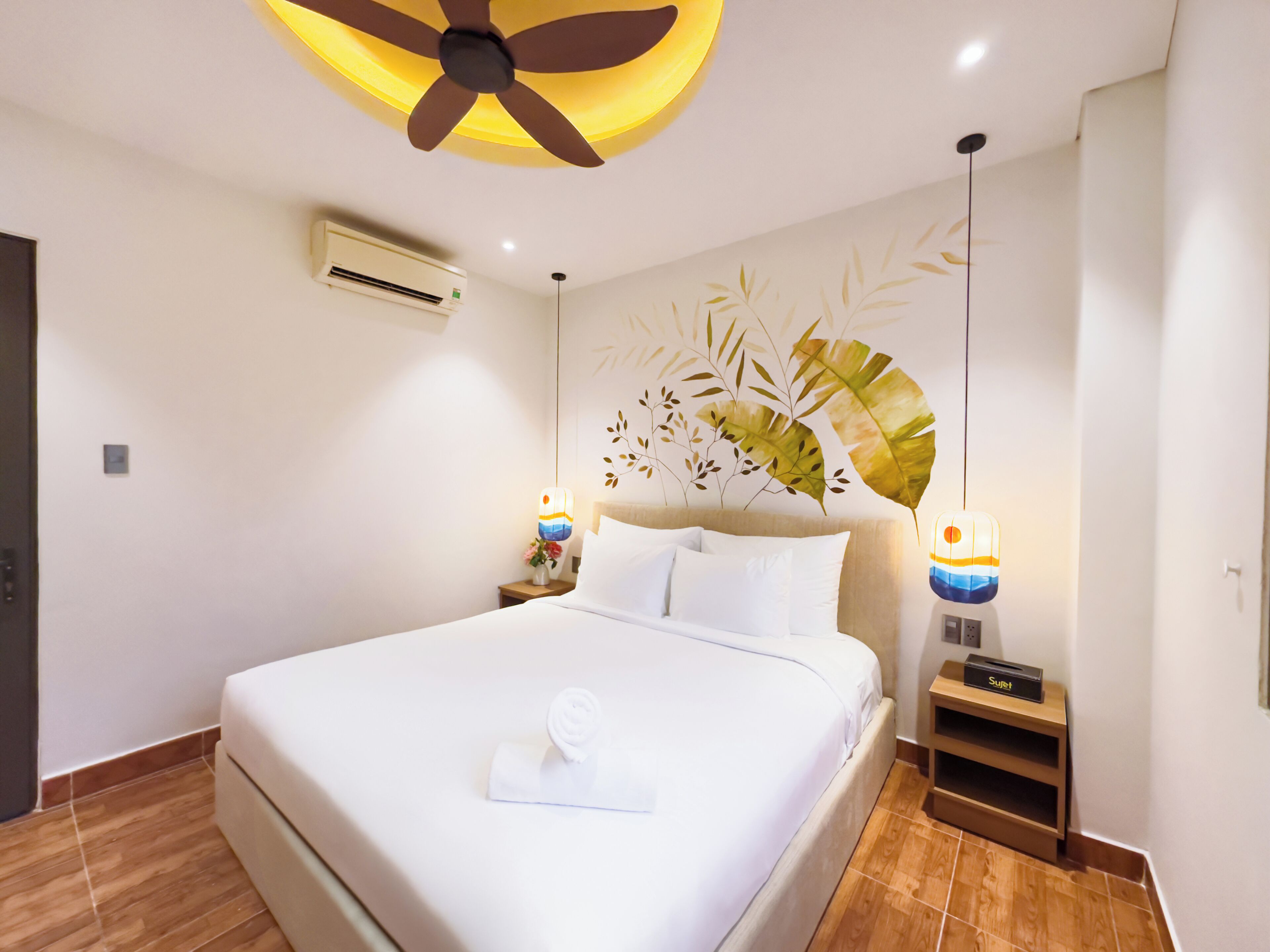 Habitación de hotel con cama, ventilador de techo, aire acondicionado de pared, un adorno en la pared y lámparas de noche.