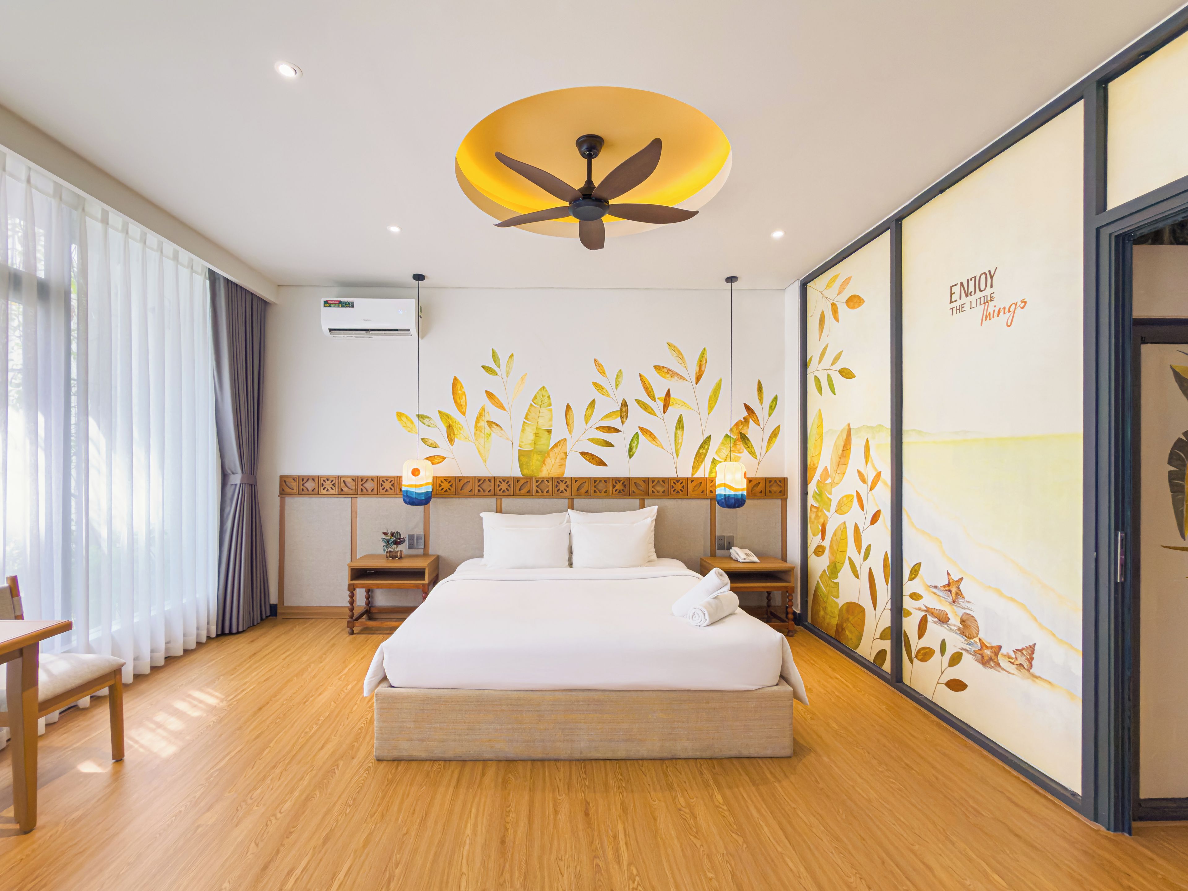 A hotel room with a large bed, wooden flooring, a ceiling fan, a wall-mounted air conditioner, and a decorative wall with leaf patterns.