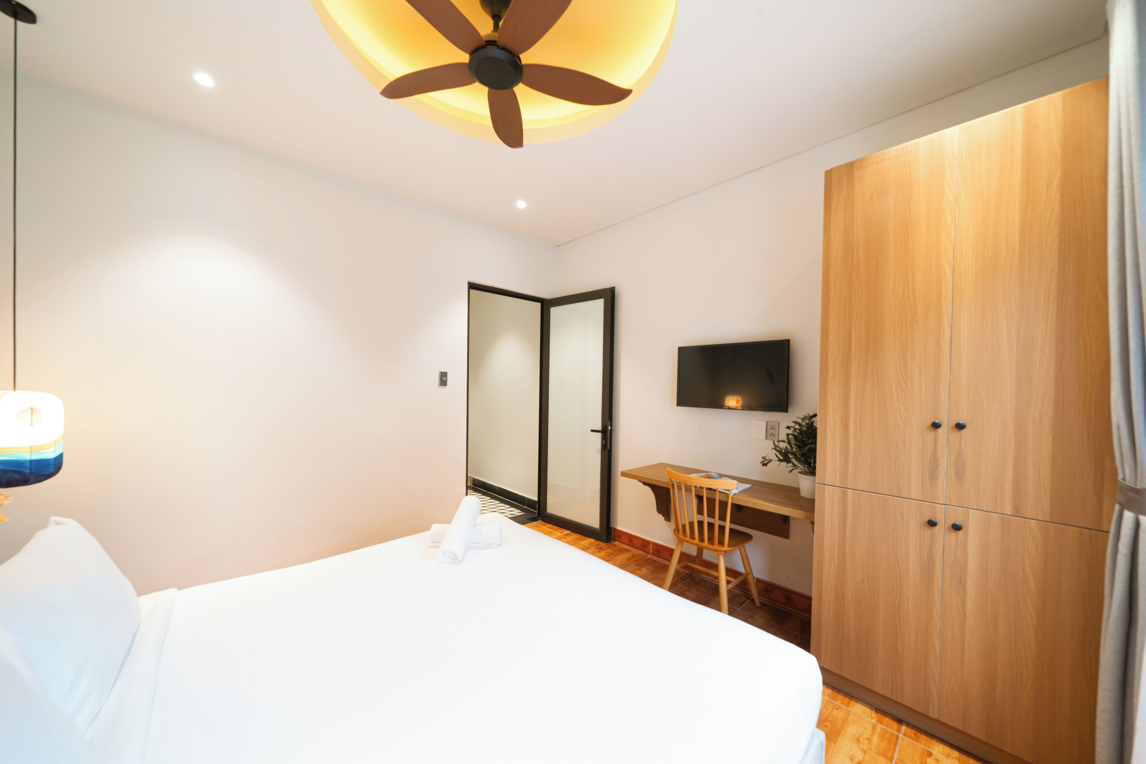A hotel room with a bed, a desk, a chair, a wardrobe, and a ceiling fan.