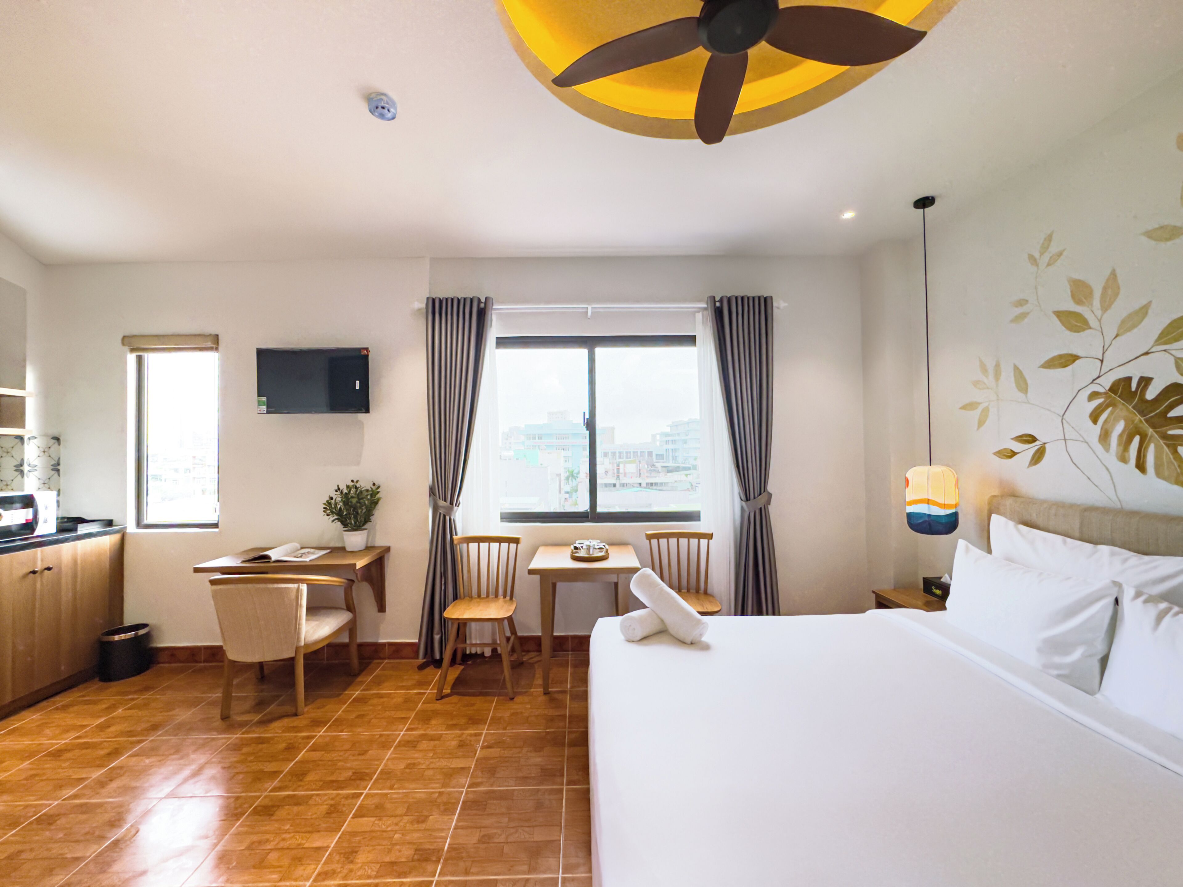 Habitación de hotel con cama, escritorio, silla, mesita, planta en maceta, televisor y ventana con cortinas.