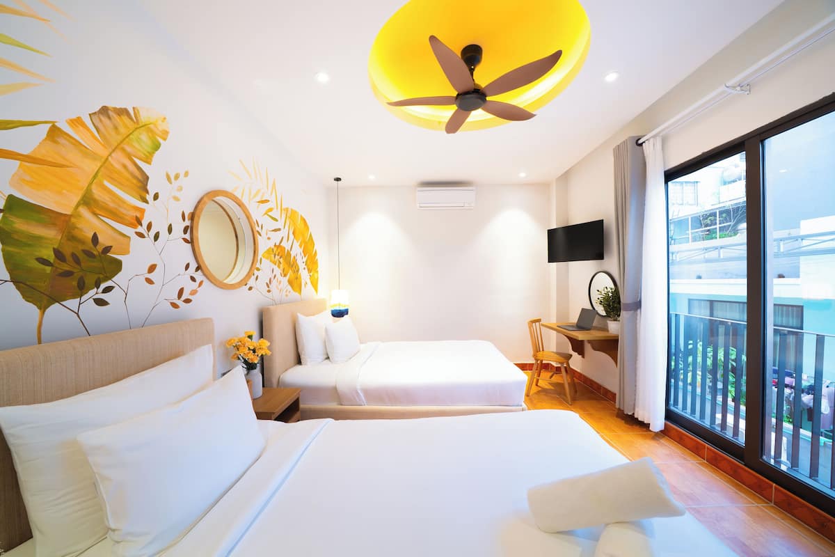 Habitación de hotel con dos camas, un ventilador de techo, un escritorio con una computadora portátil, un ventanal con vistas y una pared con un mural de hojas.