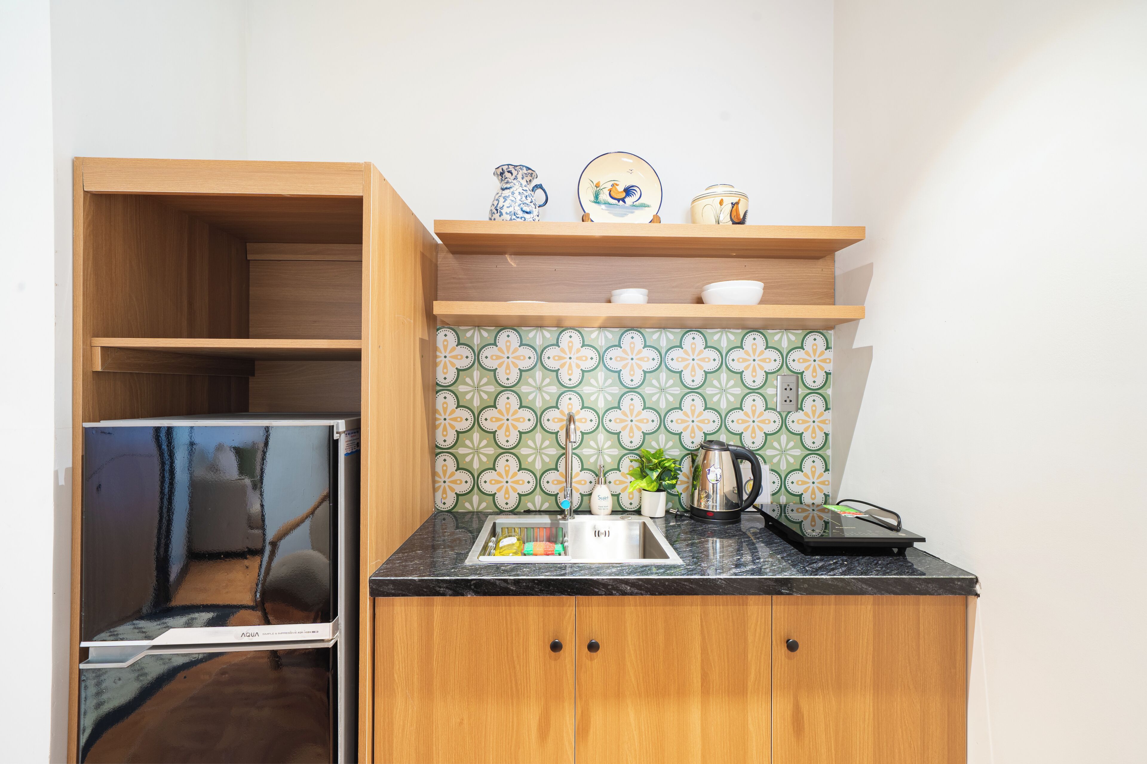 A compact kitchen with a built-in refrigerator, a sink, and a countertop. There are shelves above the sink holding decorative items and a kettle on the counter.
