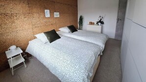 A bedroom with a textured wall, a bed with patterned bedding, a bedside table, a hanging light fixture, and a small plant on a shelf.