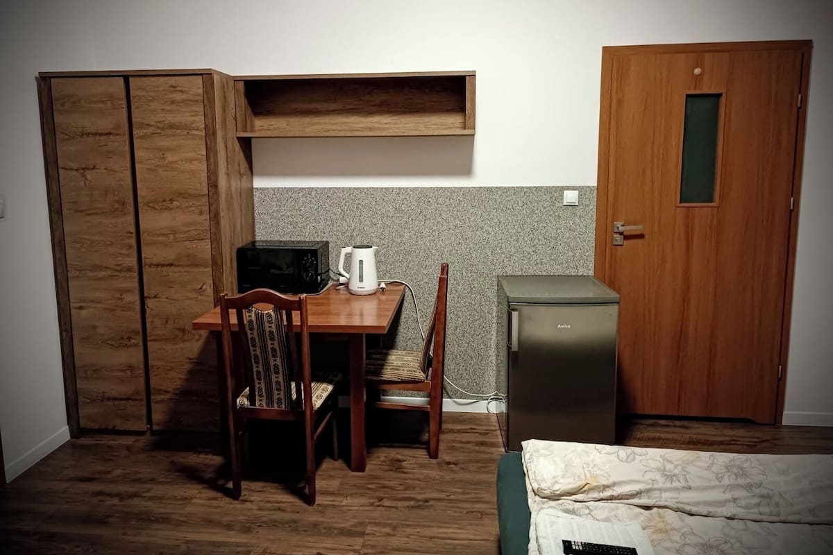 A compact room with a wooden desk, a chair, a small kitchenette, and a bed.