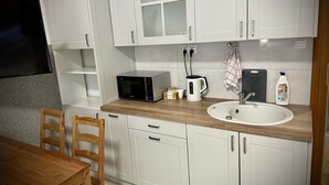 Una cocina con armarios blancos, microondas, hervidor, pileta y mesa de comedor con sillas.