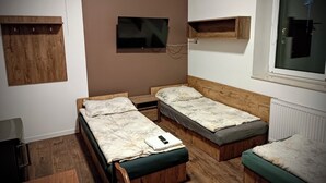Quarto pequeno e compacto com duas camas de solteiro, uma televisão na parede e uma janela com persianas.