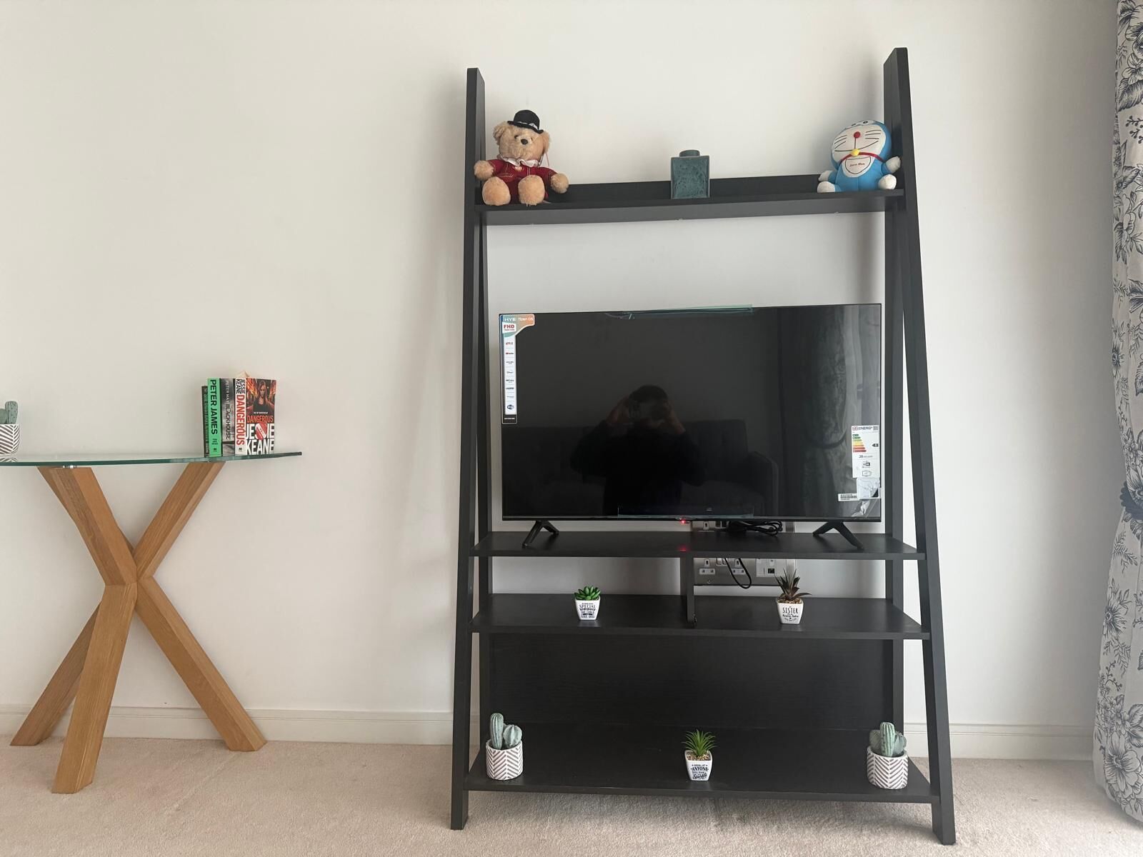 Ett svart TV-bord med en platt-TV, två hyllor med små krukväxter och ett glasbord med böcker och en dekorativ prydnad.
