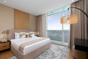 A modern hotel room with a large bed, a bedside table with a lamp, a TV, and a large window offering a city view.