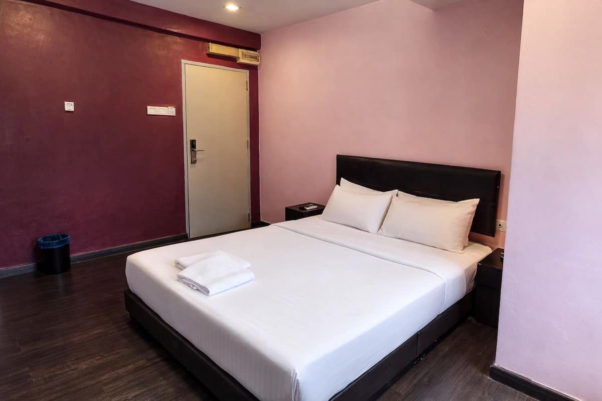 Ein ordentlich bezogenes Bett mit weißen Leinen und Kissen, ein dunkles Kopfende, ein kleiner Nachttisch mit Lampe und eine Tür mit Lichtschalter.