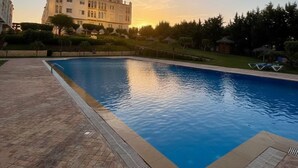 Piscina com deck revestido, rodeada por vegetação e um edifício grande ao fundo.