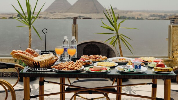 A dining table with various foods and drinks, set against a view of the pyramids. - deluxe double room with mountain view (Al Haram Cairo)