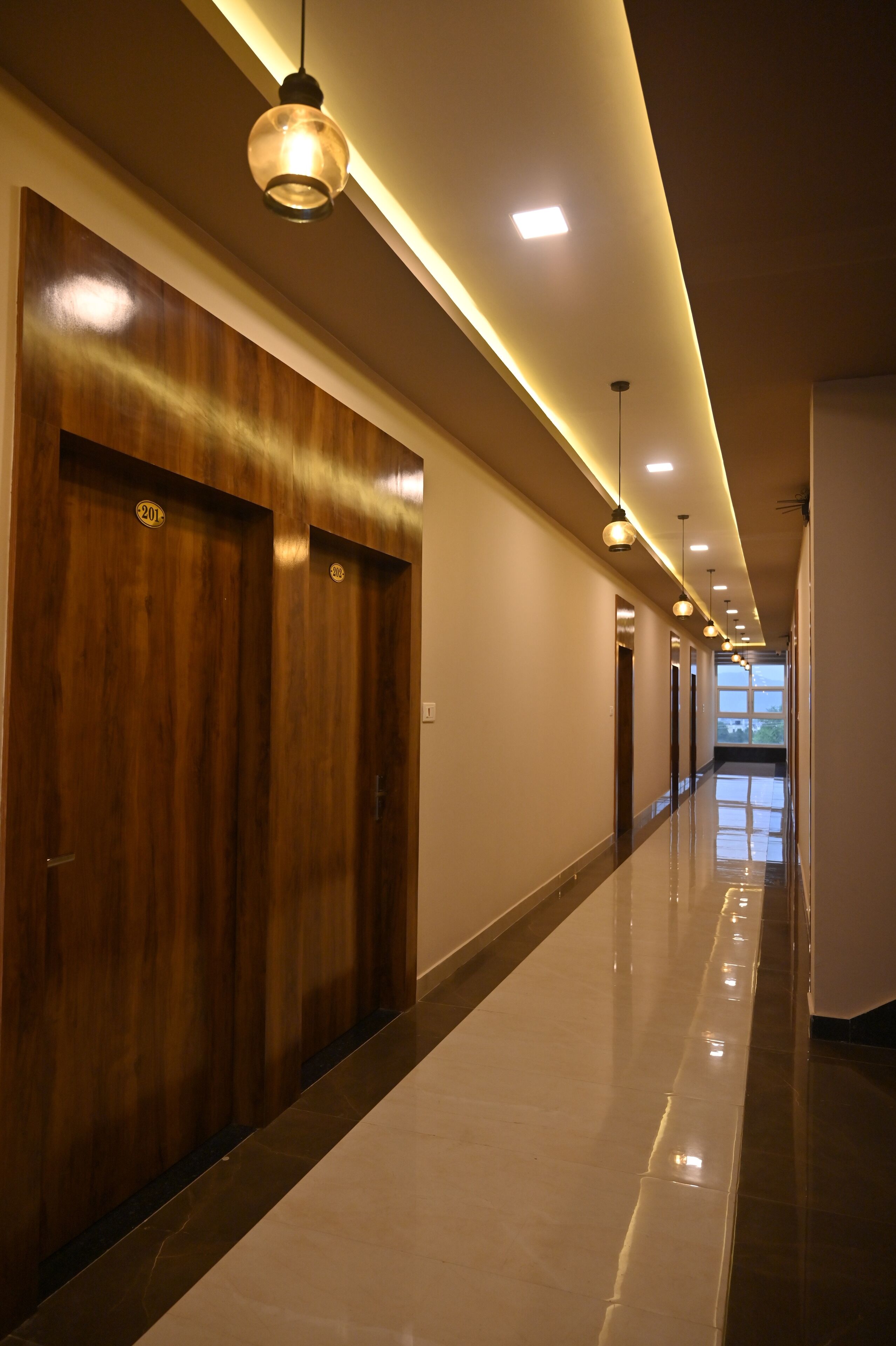 A long, well-lit hallway with wooden doors and a shiny floor, leading to a bright room with a large window.