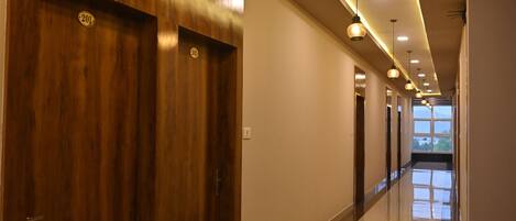 A long, well-lit hallway with wooden doors and a shiny floor, leading to a bright room with a large window.