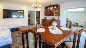 Un comedor con mesa de madera, sillas y una araña. Se observa una cocina al fondo con armarios de madera y una ventana.