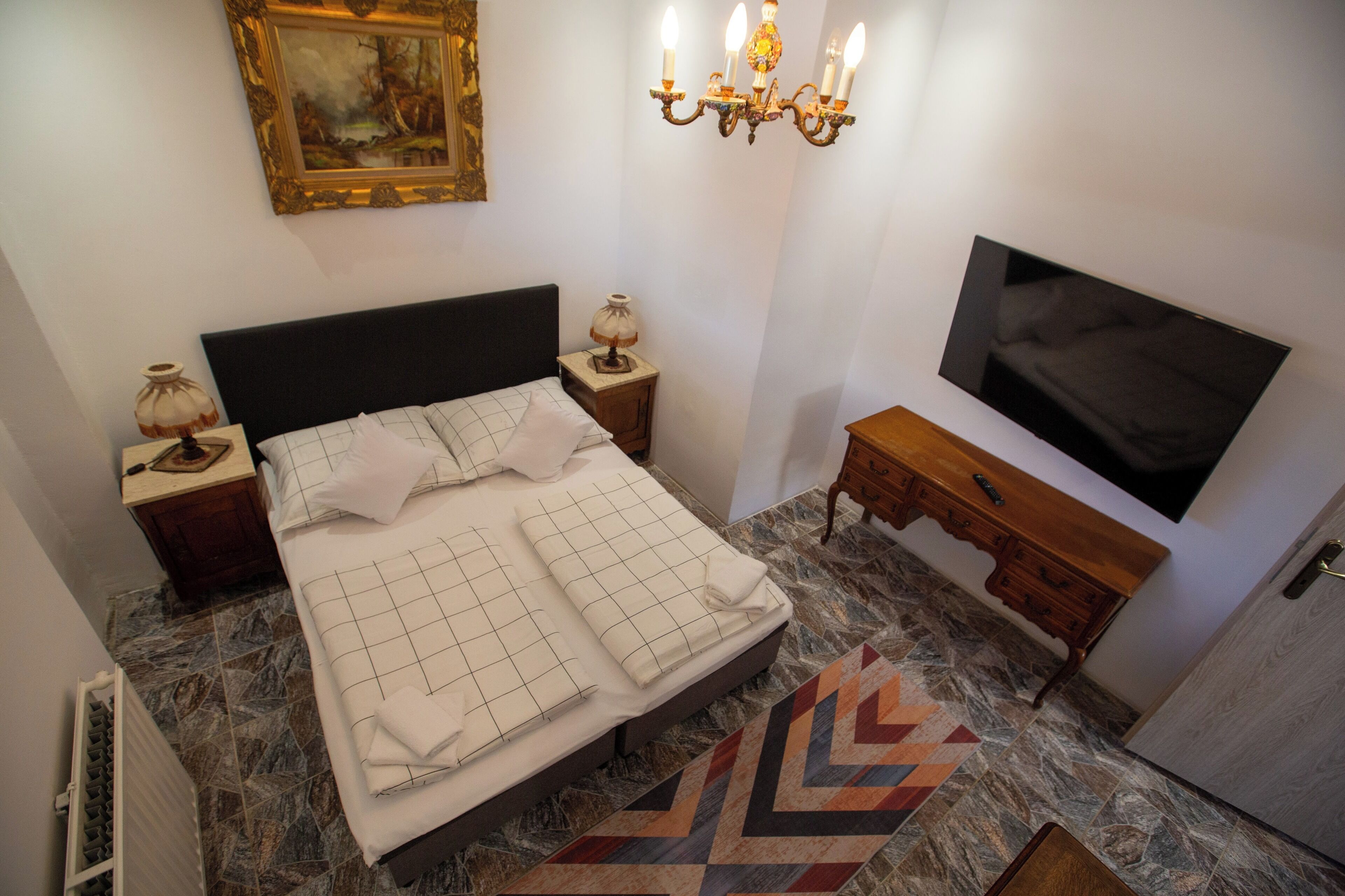 Quarto com cama, mesas de cabeceira, televisão, lustre e um quadro na parede.
