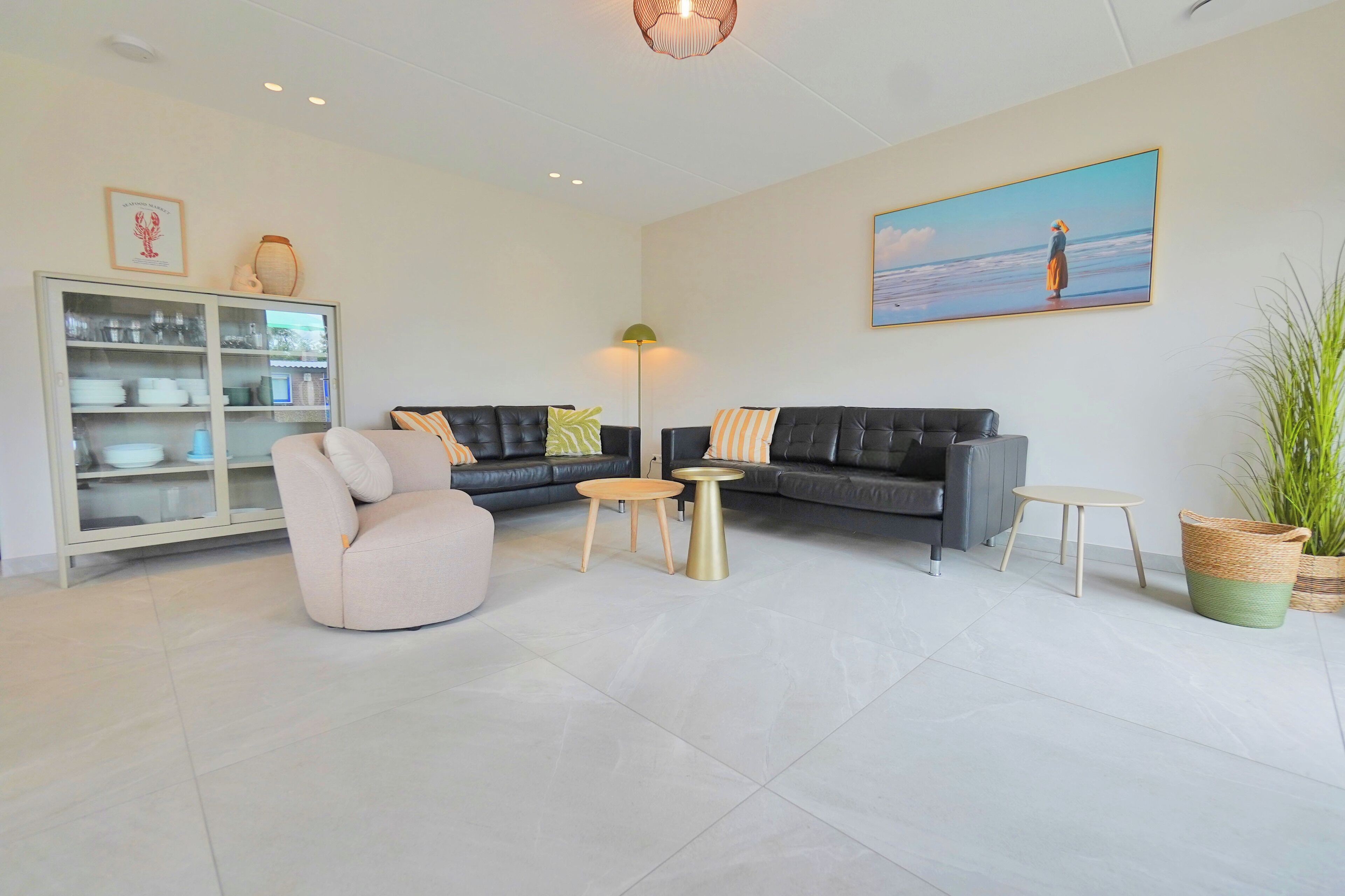A modern living room with a sofa, armchair, and coffee table, a cabinet with dishes, and a painting of a beach scene.