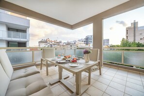 A balcony with a dining table set for four, chairs, and a sofa. The balcony overlooks a cityscape with buildings and a green area. - Lena Court - Spacious 3bdr, City Views (Limassol)