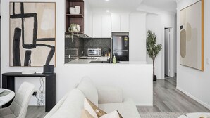 A modern kitchen with white cabinets, a black refrigerator, and a microwave. There is a dining area with a white table and chairs, and a living room with a sofa and a coffee table. - Modern City Escape | Stylish 2BR Apartment (East Perth)