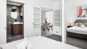 A modern hotel room with a large bed, a bedside table with a lamp, a bathroom with a mirror and sink, and a dining area with a table and chairs. - Modern City Escape | Stylish 2BR Apartment (East Perth)