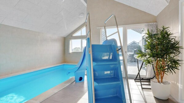 An indoor swimming pool with a blue slide, a potted plant, and a view of the outside.