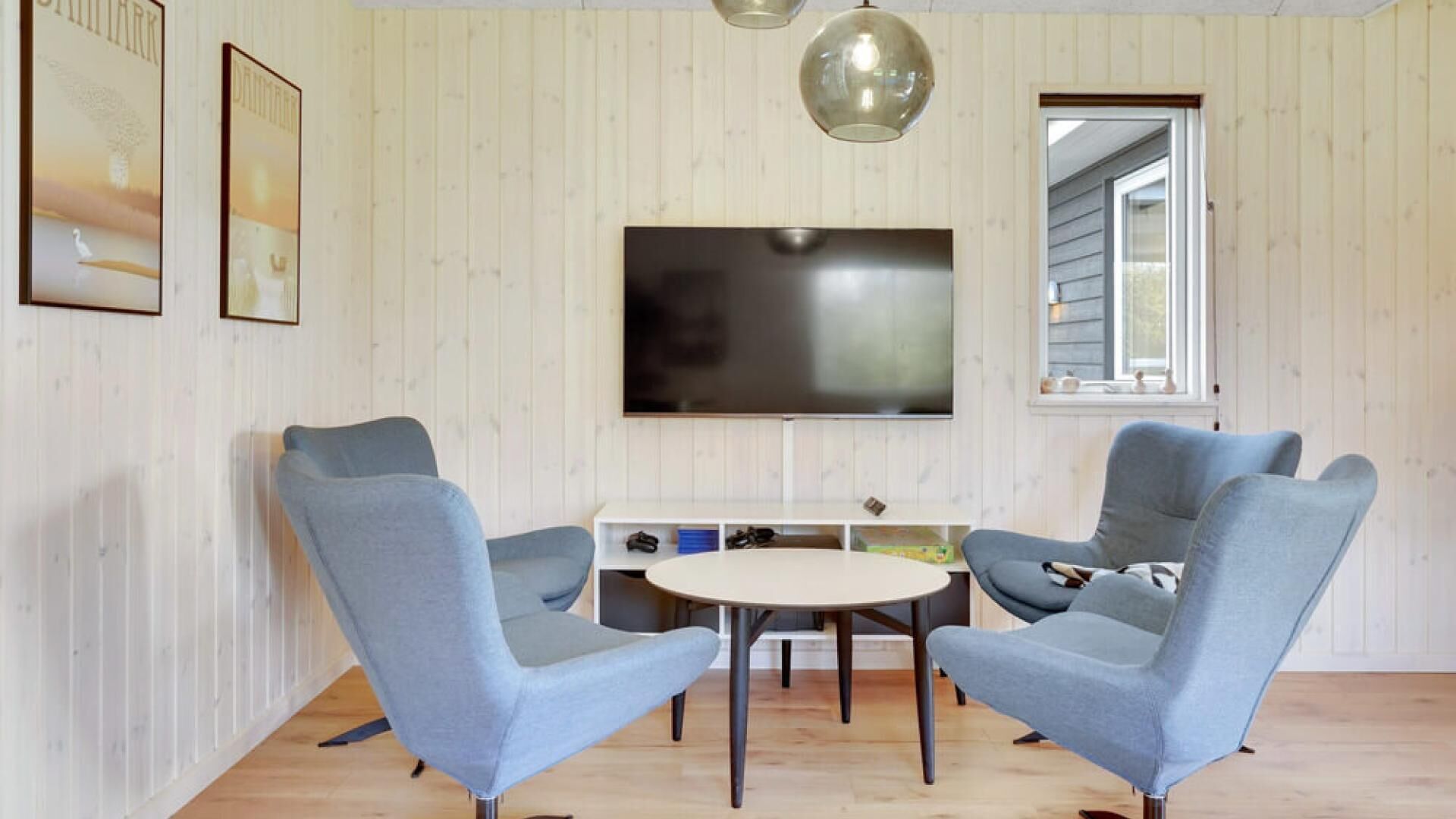 En moderne stue med to blå lænestole, et rundt bord, et fladskærms-tv ophængt på væggen og et vindue med udsigt.