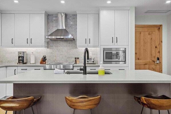 A modern kitchen with white cabinets, a stainless steel range hood, a built-in microwave, and a large island with a sink. There are three wooden bar stools in front of the island. - Creekside Cove Lux 1Bd close to DT w hot tub (Canmore)