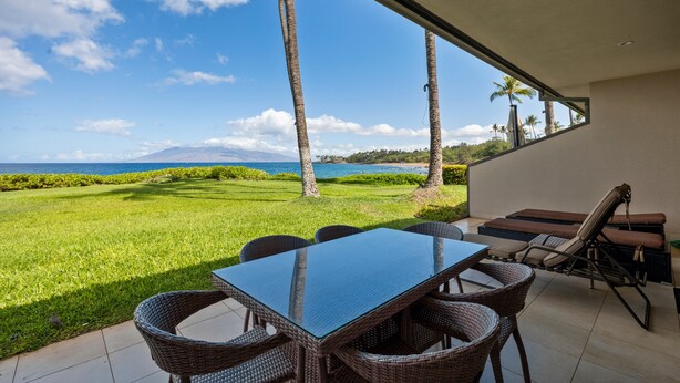 Um pátio com mesa e cadeiras, com vista para uma área gramada com palmeiras e o oceano.