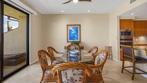 Uma sala de jantar com uma mesa de vidro redonda, rodeada por cadeiras de vime, um ventilador de teto e um quadro pendurado na parede.