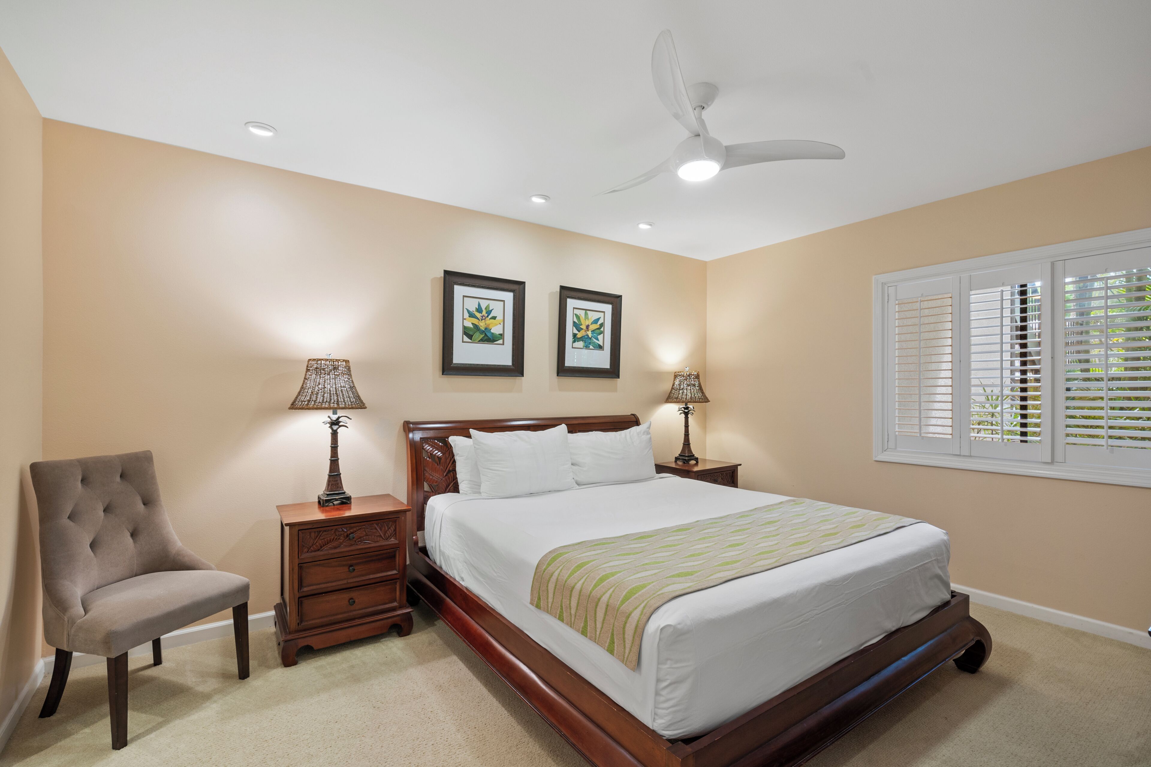 Une chambre à coucher comprenant un lit, une chaise, deux tables de chevet avec des lampes, un ventilateur de plafond et une fenêtre avec des stores.