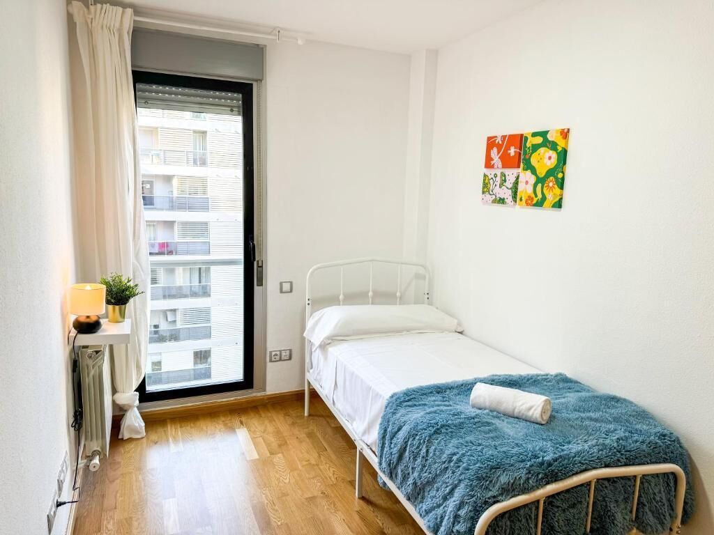A small, single-bed room with a wooden floor, a window with a view of buildings, a radiator, a bedside table with a lamp, and a colorful artwork on the wall.