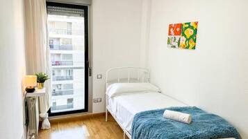 A small, single-bed room with a wooden floor, a window with a view of buildings, a radiator, a bedside table with a lamp, and a colorful artwork on the wall.
