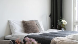 Een netjes opgemaakt bed met kussens, een nachtkastje, een vaas met bloemen en een raam met gordijnen.