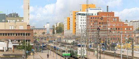 A train station with multiple tracks, a green train, and surrounding buildings including a tall tower and a building with 'TECHNOPOLIS' signage.