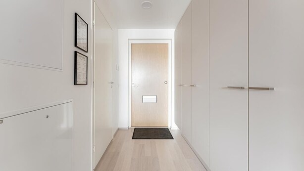 A narrow hallway with white walls, a wooden door at the end, and a black doormat on the floor. - Modern studio with own glazed balcony for 3 people (Tampere)