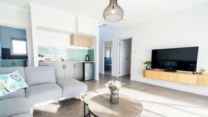 A modern living room with a grey sofa, a round coffee table with a vase, a flat-screen TV, and a kitchen in the background. - Helios Heights - Modern 2 storey, Off Peak Special available (Moonta Bay)