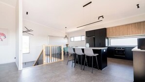 A modern kitchen with a central island, bar stools, and a dining area with a table and chairs. - Helios Heights - Modern 2 storey, Off Peak Special available (Moonta Bay)