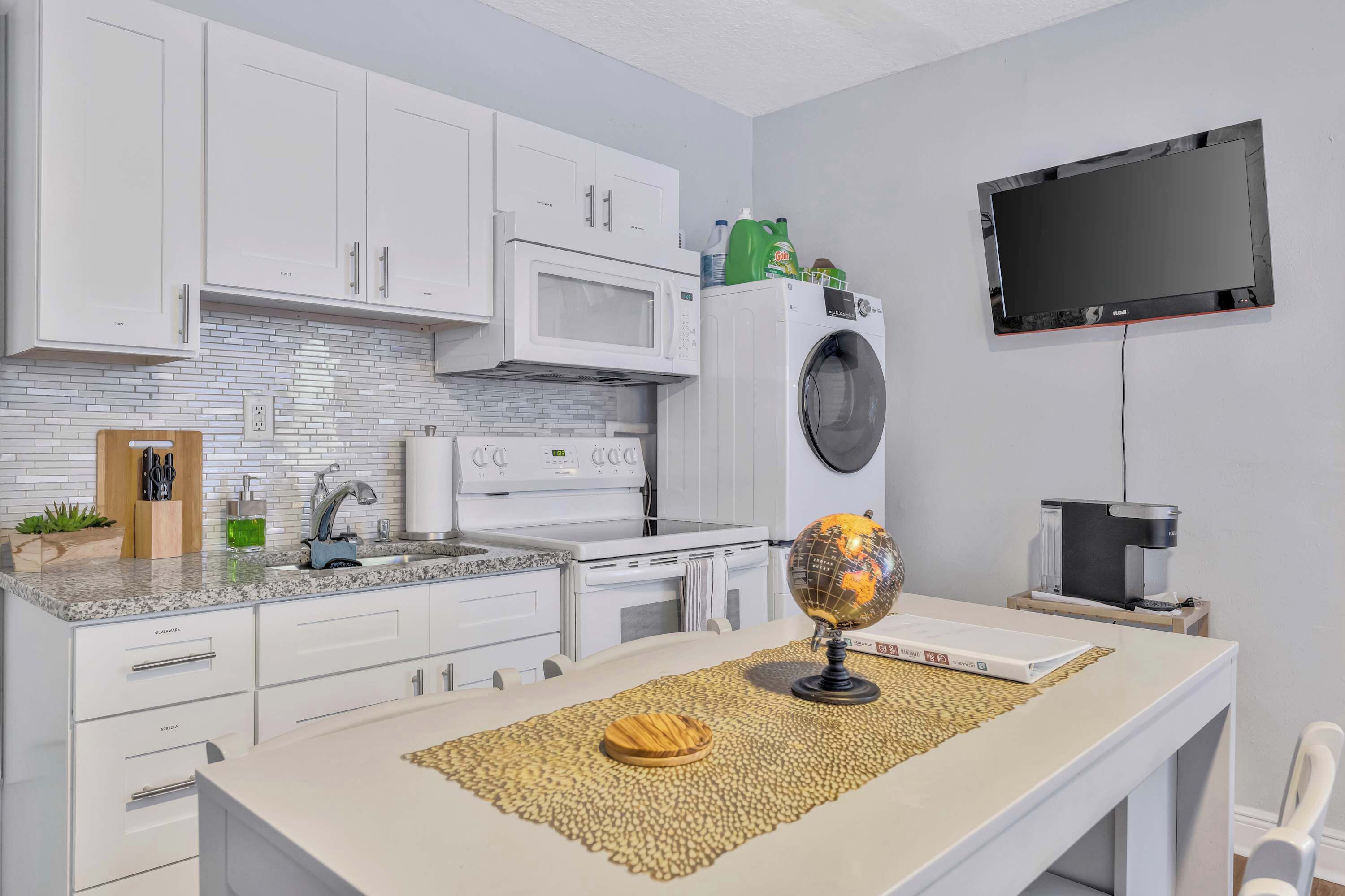 Een moderne keuken met witte kastjes, een magnetron, een wasmachine en een flatscreen televisie.