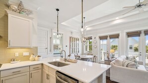 A modern kitchen with white cabinetry, a dishwasher, and a large island with a sink. There is a dining area with a table and chairs, and a living room with a sofa and a ceiling fan. - ☀️ Sun & Sand for 6! NEW 6 Seater Golf Cart | Heated Pool | 4 Bikes 🚲 (Santa Rosa Beach)