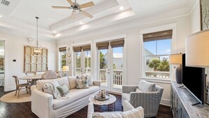 A spacious living room with a white sofa, armchairs, and a coffee table. There is a ceiling fan, a chandelier, and large windows with blinds. - ☀️ Sun & Sand for 6! NEW 6 Seater Golf Cart | Heated Pool | 4 Bikes 🚲 (Santa Rosa Beach)