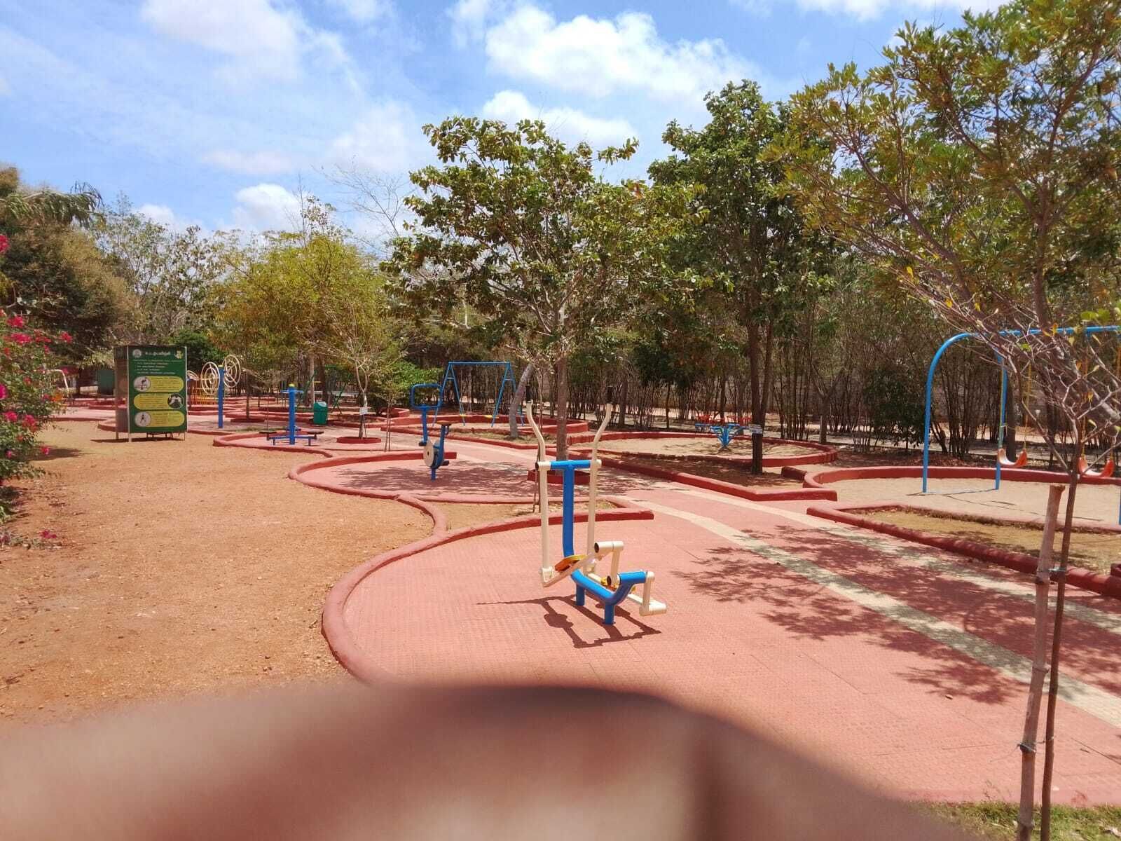 Un espacio al aire libre para ejercitarse con diversos equipos de gimnasia, rodeado de árboles y un sendero pavimentado.