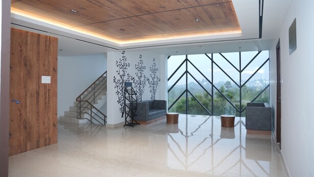 A spacious, modern interior with a wooden ceiling, a staircase, a grey sofa, and a large window offering a view of the outdoors. - Vanaalayam Retreat Palladam by Arcadia (Palladam)