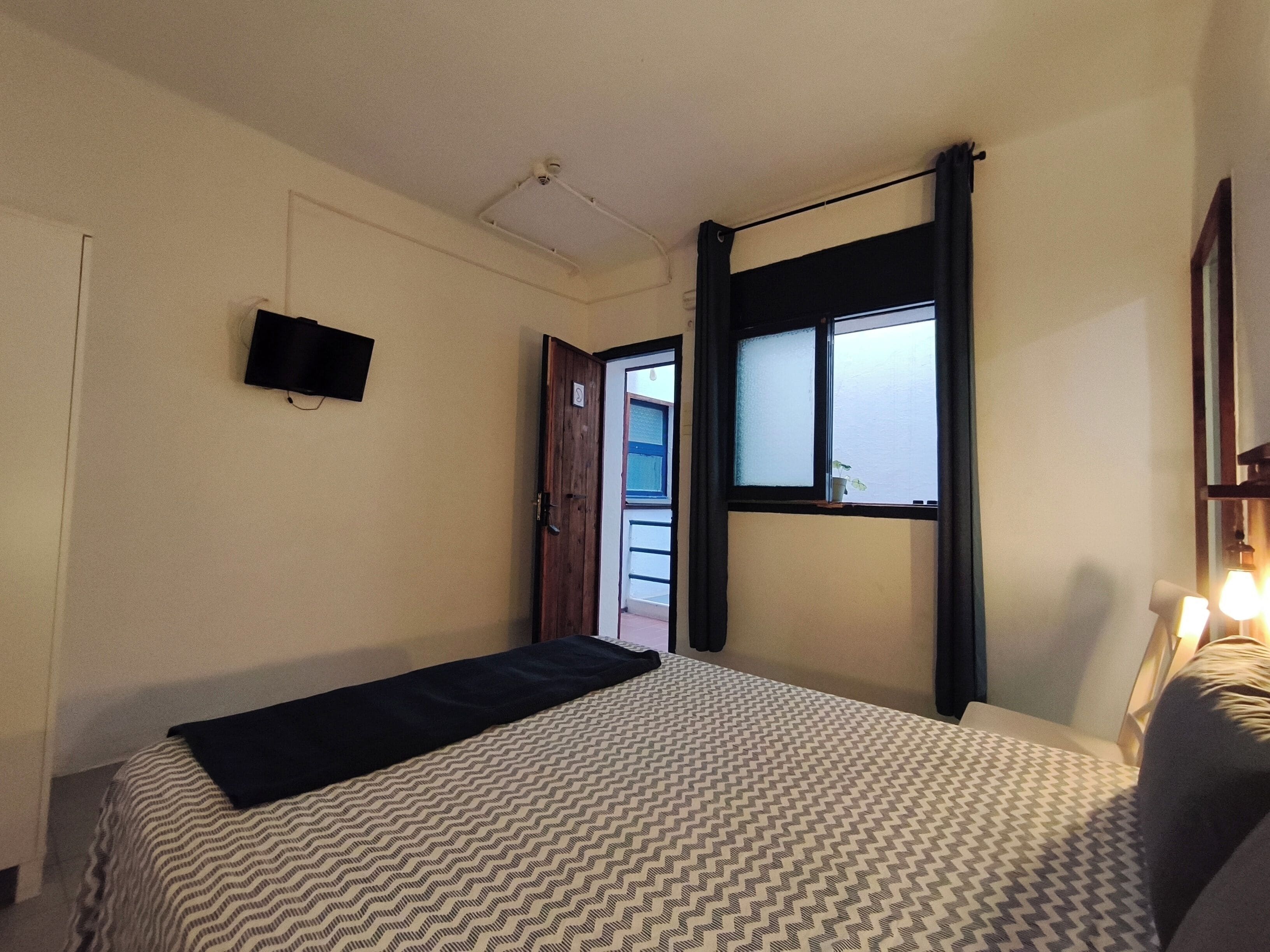 Un dormitorio con una cama, un televisor montado en la pared, un ventanal con cortinas y una puerta que da a otra habitación.