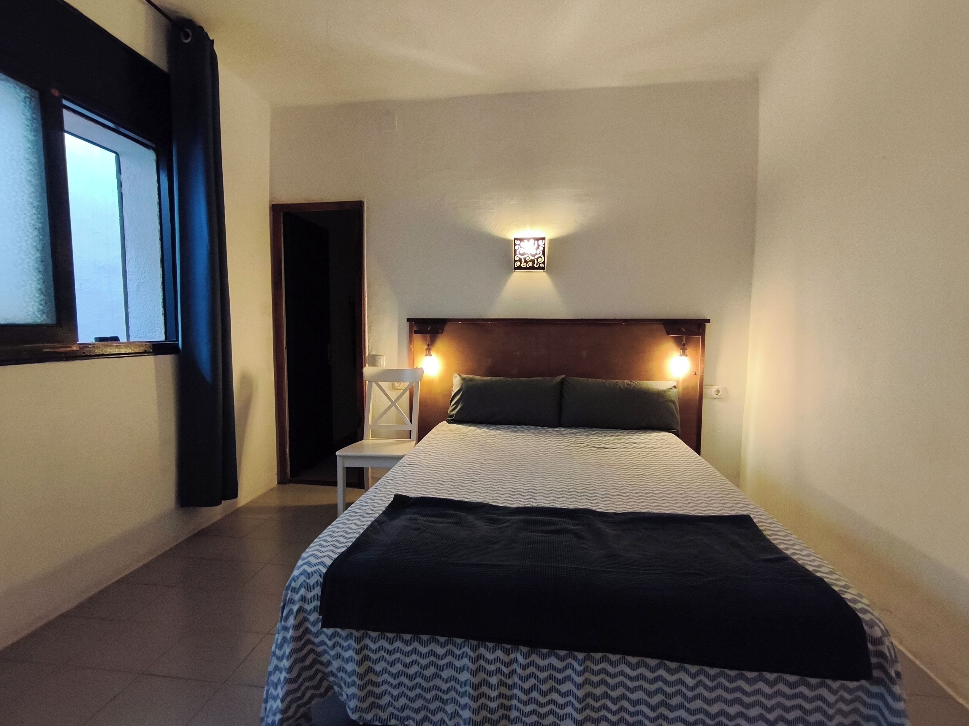A hotel room with a bed, a nightstand, a wall-mounted lamp, and a window with curtains.