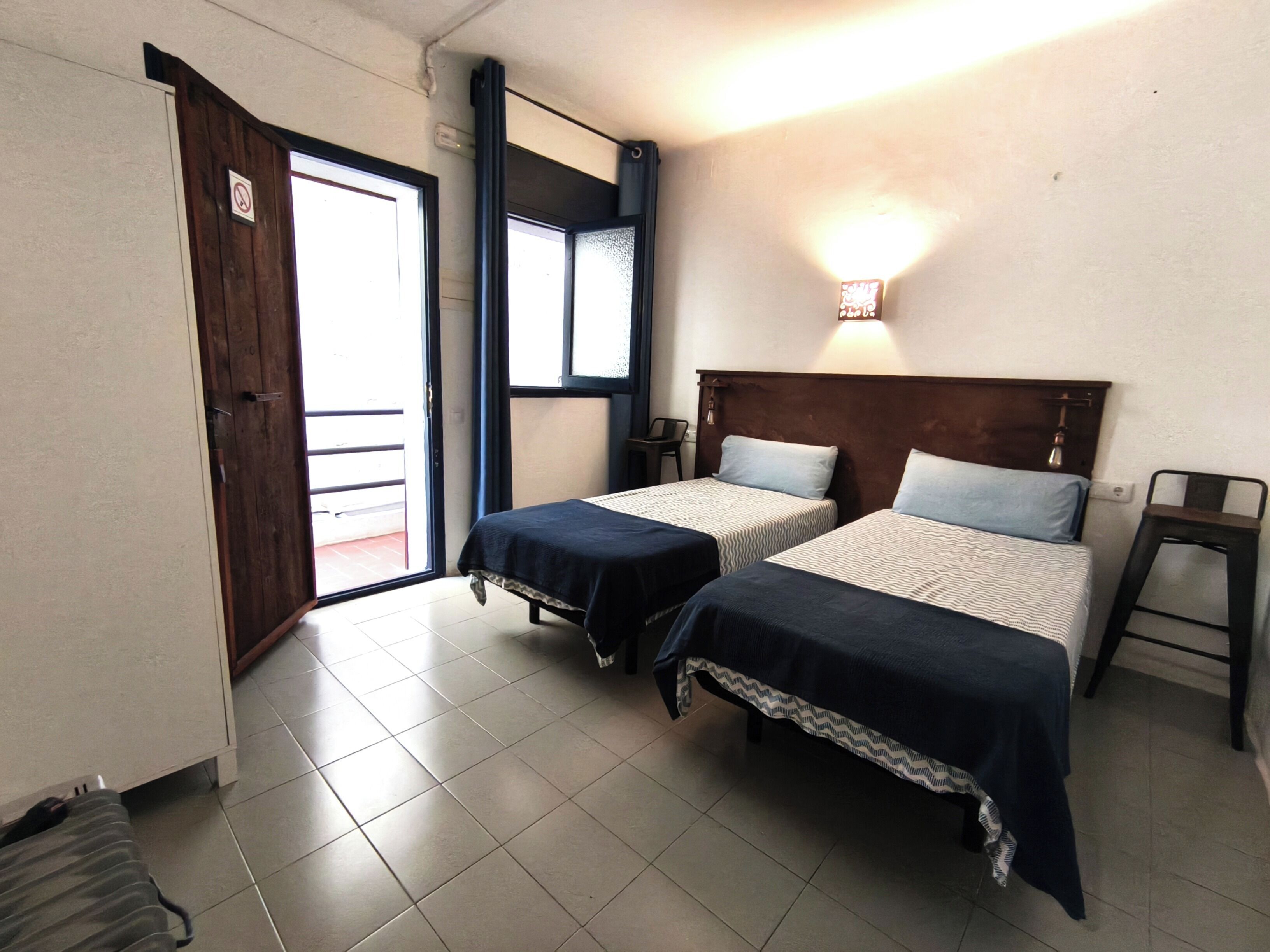 A room with two beds, a wooden door, a window, and a wall-mounted light.